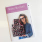 ront cover of a printed sewing pattern booklet titled "Baez Bucket" by Natalie Santini (@sewhungryhippie). The cover features a purple banner at the top and a photo of a smiling woman with glasses, a leopard print headband, and a denim shirt. She is holding a bucket-style fabric tote bag with a dark, patterned floral design and a pink fabric lining.
