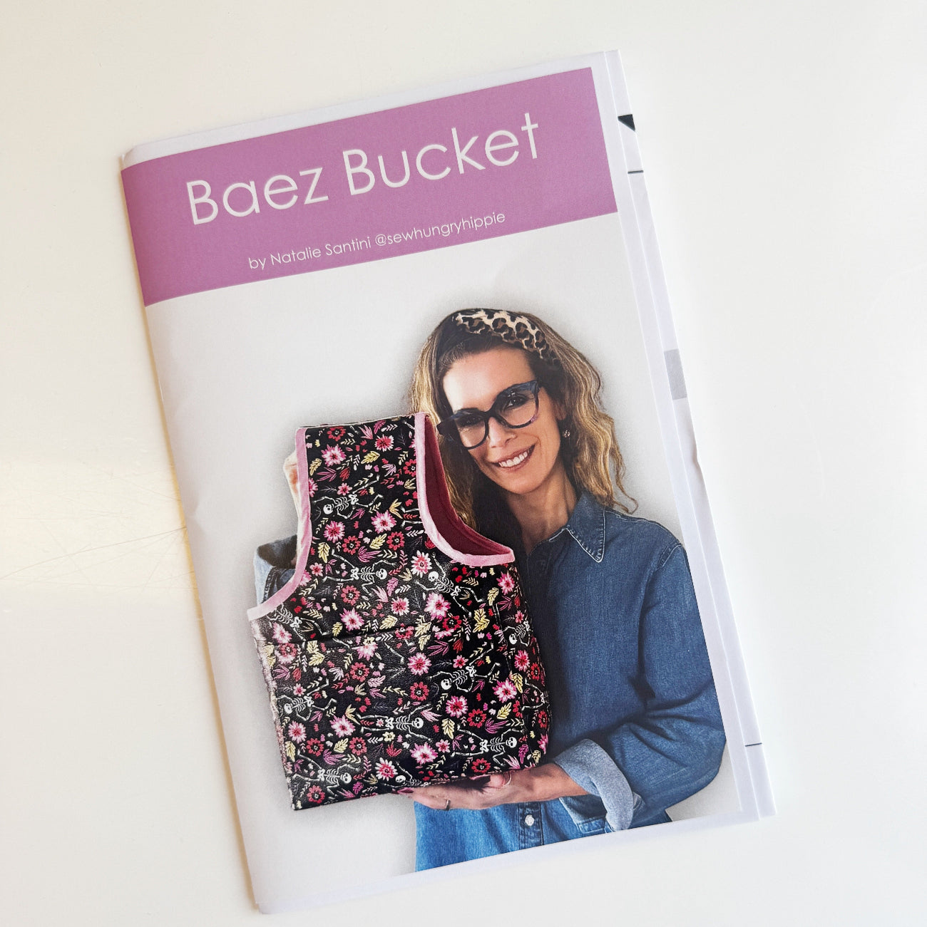 ront cover of a printed sewing pattern booklet titled "Baez Bucket" by Natalie Santini (@sewhungryhippie). The cover features a purple banner at the top and a photo of a smiling woman with glasses, a leopard print headband, and a denim shirt. She is holding a bucket-style fabric tote bag with a dark, patterned floral design and a pink fabric lining.