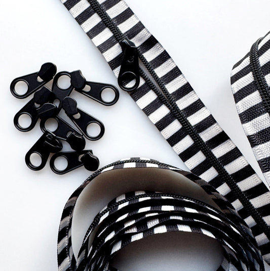 Black and white striped zipper ribbon with black metal zipper teeth, shown coiled on a white background with several loose zipper teeth pieces.