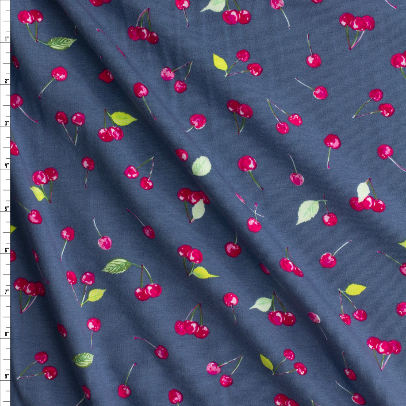 A close-up of draped fabric featuring a repeating cherry print on a slate blue-gray background. The pattern includes bright red cherries with green and yellow leaves and stems.