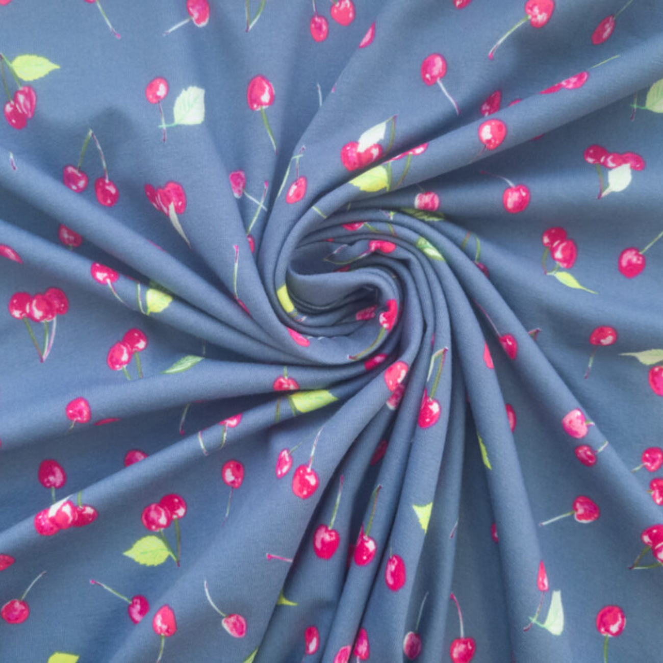 A close-up of blue-gray fabric arranged in a spiral, printed with a repeating pattern of bright red cherries and small green leaves.