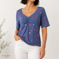 A woman wearing a short-sleeve V-neck top with a gathered front and a blue cherry print, paired with white pants, against a light background.