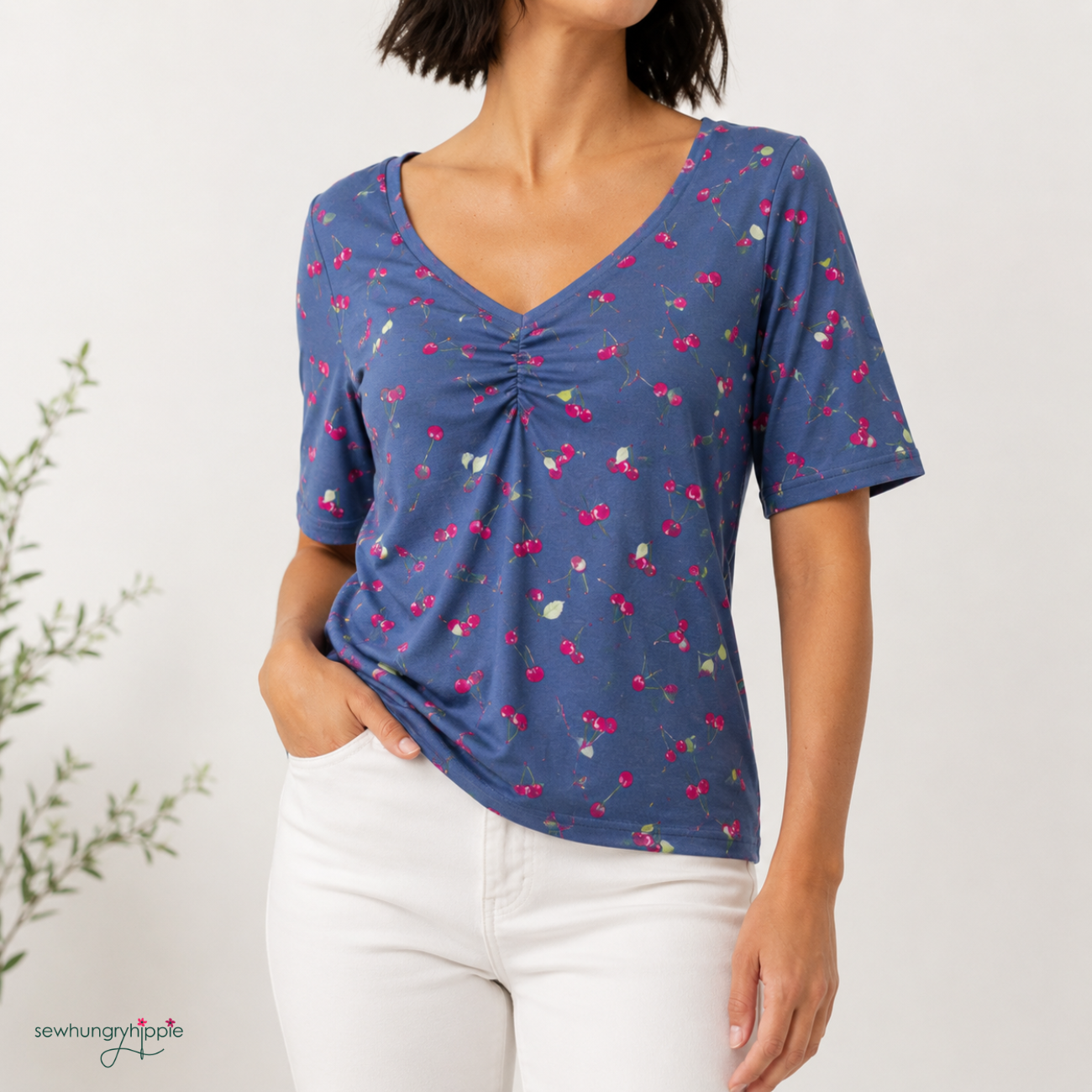 A woman wearing a short-sleeve V-neck top with a gathered front and a blue cherry print, paired with white pants, against a light background.
