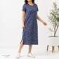 A woman models a short-sleeve midi dress with a small pink and yellow cherry print on a navy blue background. She is wearing white sneakers and standing in a bright room next to a potted plant.