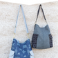 Two handbags, one blue with geometric pattern and one gray with black accents, hanging against a textured wall. Pattern name 'Compass Bag' and brand 'noodlehead' visible.