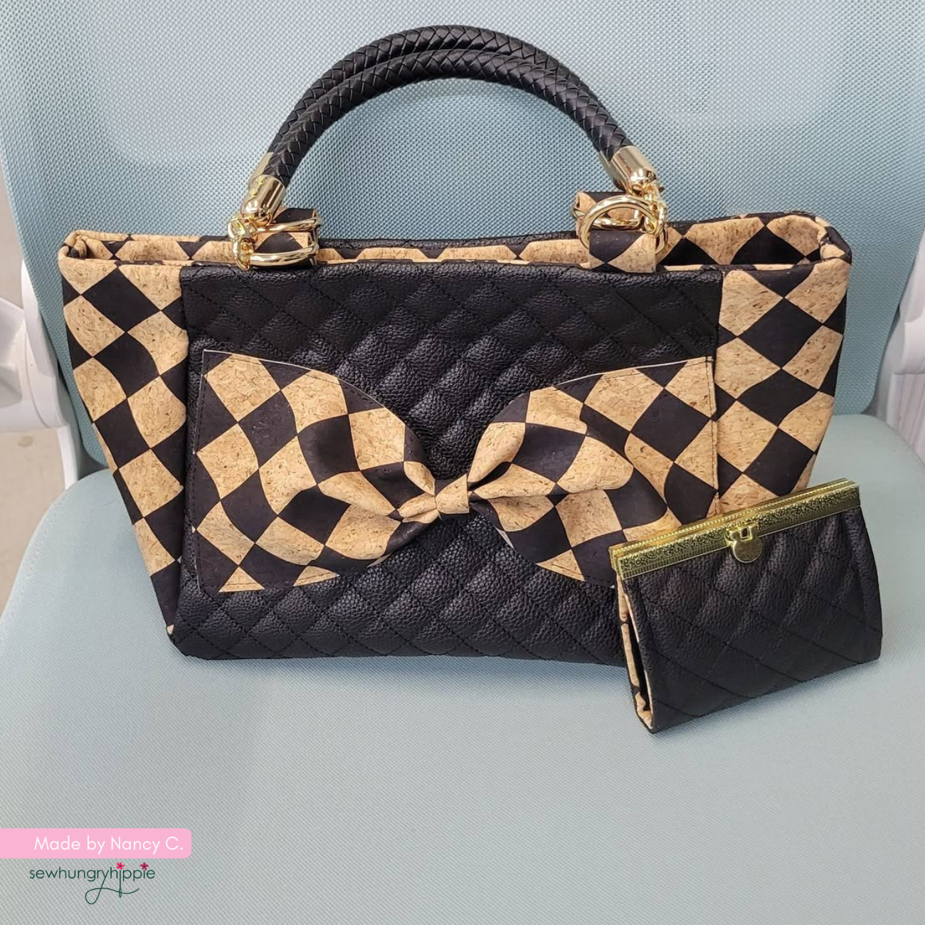 A black quilted handbag with a tan and black checkered pattern on the trim, handle, and front pocket, accompanied by a matching small wallet. The bag features a black braided top handle and gold-tone hardware, displayed on a light blue chair.