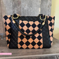 A structured tote bag with a black and tan checkered pattern made from quilted leather and cork panels, featuring a black braided shoulder strap and gold-tone hardware, displayed on a rustic wooden surface.