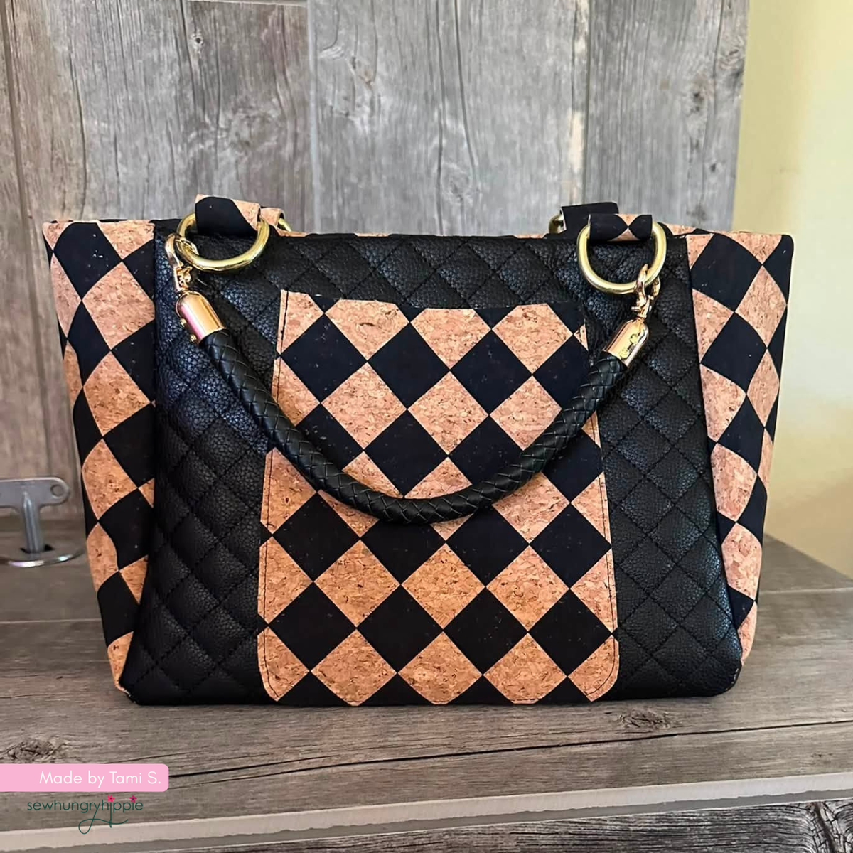 A structured tote bag with a black and tan checkered pattern made from quilted leather and cork panels, featuring a black braided shoulder strap and gold-tone hardware, displayed on a rustic wooden surface.