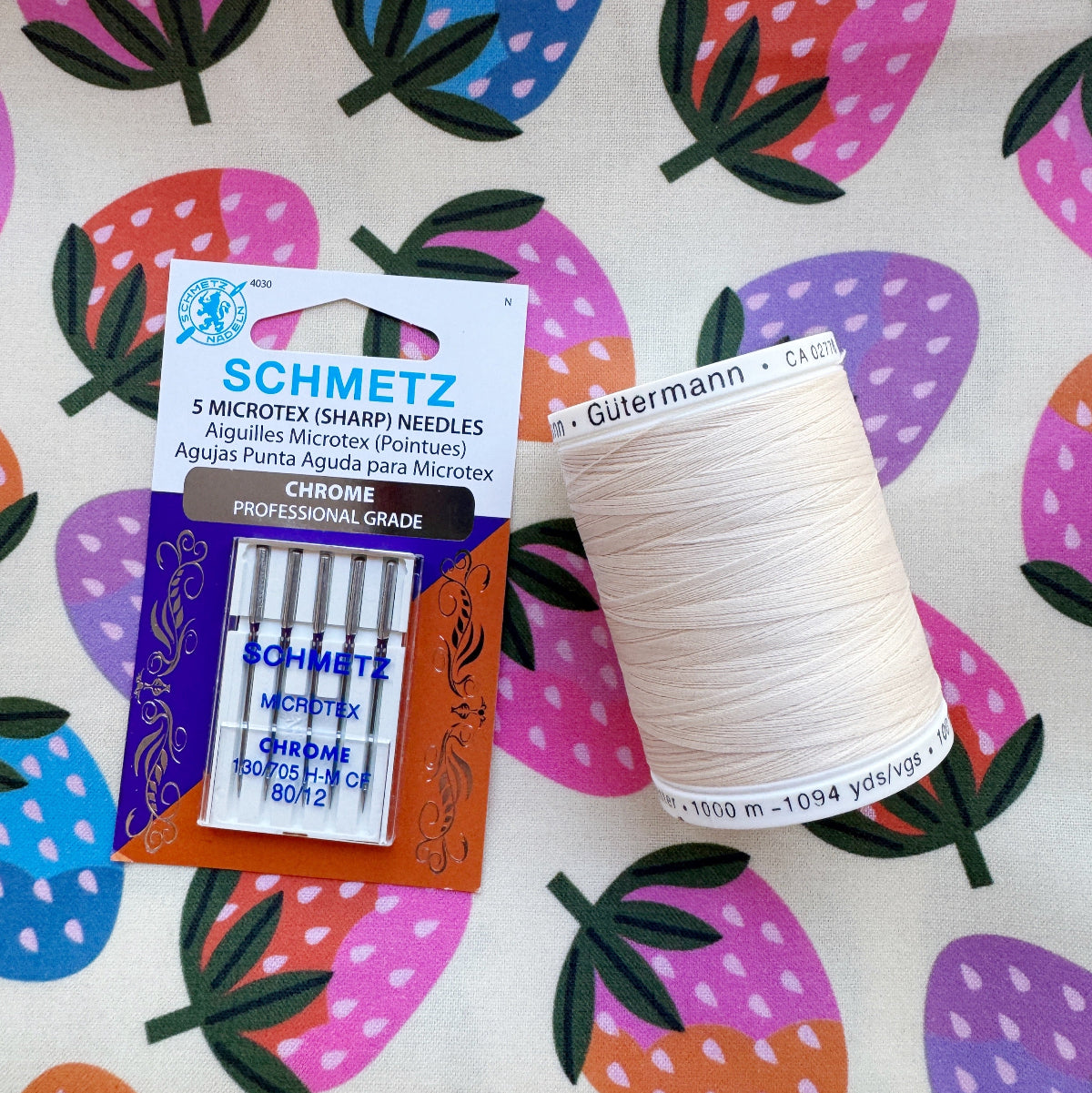 A package of Schmetz 5 Microtex (Sharp) Chromе sewing needles next to a spool of Gutermann thread, placed on a strawberry-patterned fabric background.