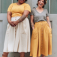 Two women standing together wearing floral crowns, yellow and white skirts, and brown boots.
