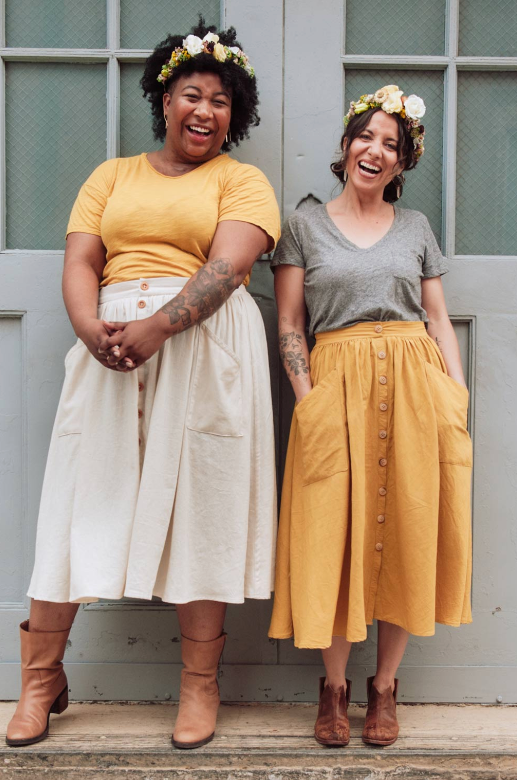 Two women standing together wearing floral crowns, yellow and white skirts, and brown boots.