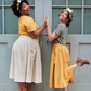 Two women standing on a step, wearing matching outfits with yellow and gray shirts and beige skirts.