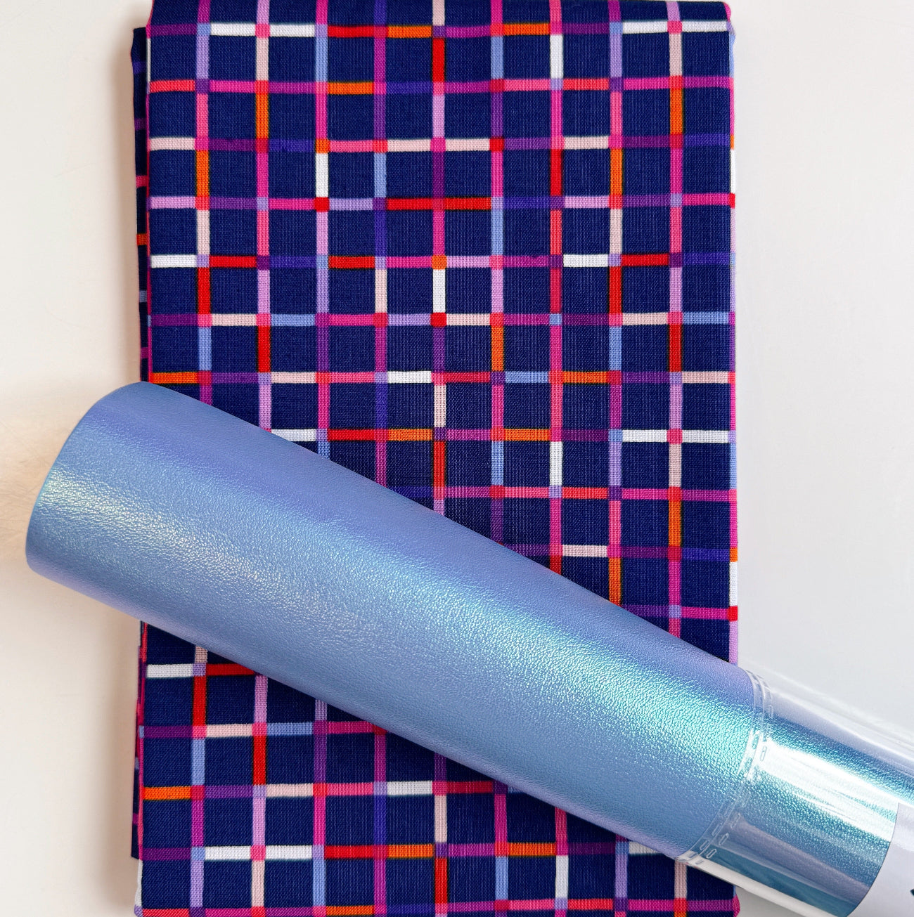 Dark navy blue cotton fabric with a colorful geometric grid plaid pattern paired with a coordinating iridescent light blue faux leather vinyl roll for handmade bag making and sewing.