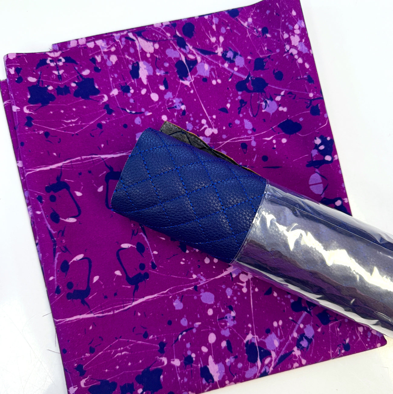 A flat-lay view of the craft materials for the Match Up #6 package, showing a piece of purple fabric with a splatter paint design next to a roll of blue pre-quilted, pebbled vinyl.