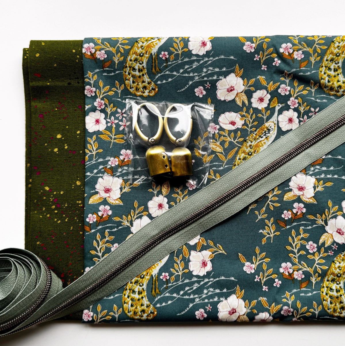 Sewing and bag crafting kit featuring a teal fabric with a peacock and floral pattern, paired with green speckled red yellow accent fabric, a zipper sage green, and bronze tassle hardware.