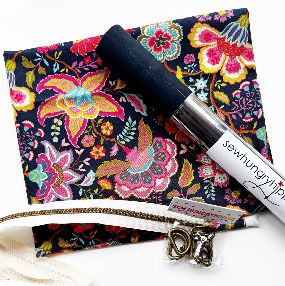 Sewing kit or bag-making supply bundle from Sew Hungryhippie, featuring a dark floral print fabric, a roll of navy blue cork  a white zipper tape in bronze teeth, and  bronze hardware d-rings and shivels.