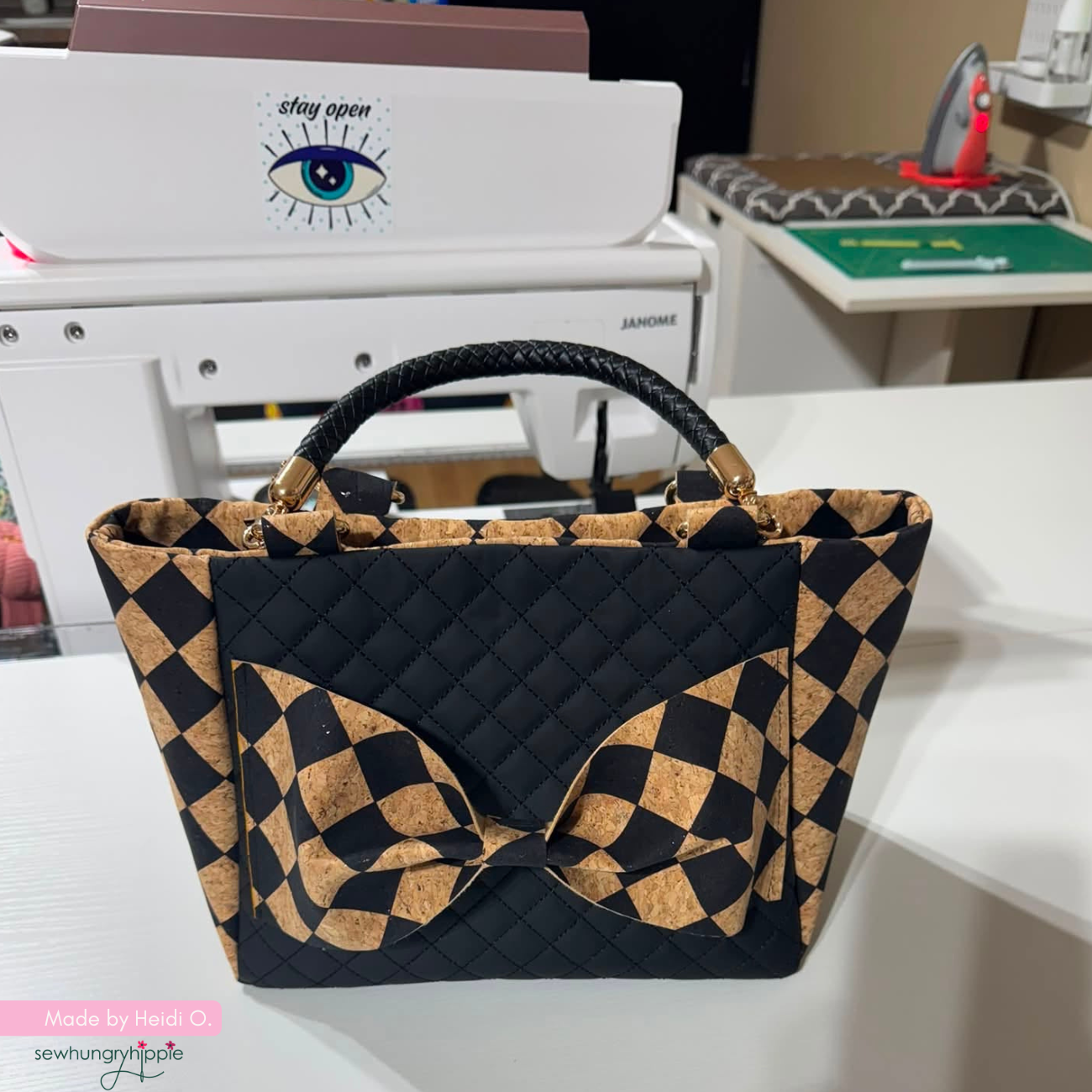 A handmade tote bag with a black and tan checkerboard pattern, featuring a quilted black front panel and a large matching bow on the front. The bag is placed on a white table with a sewing machine and other workshop items in the background.