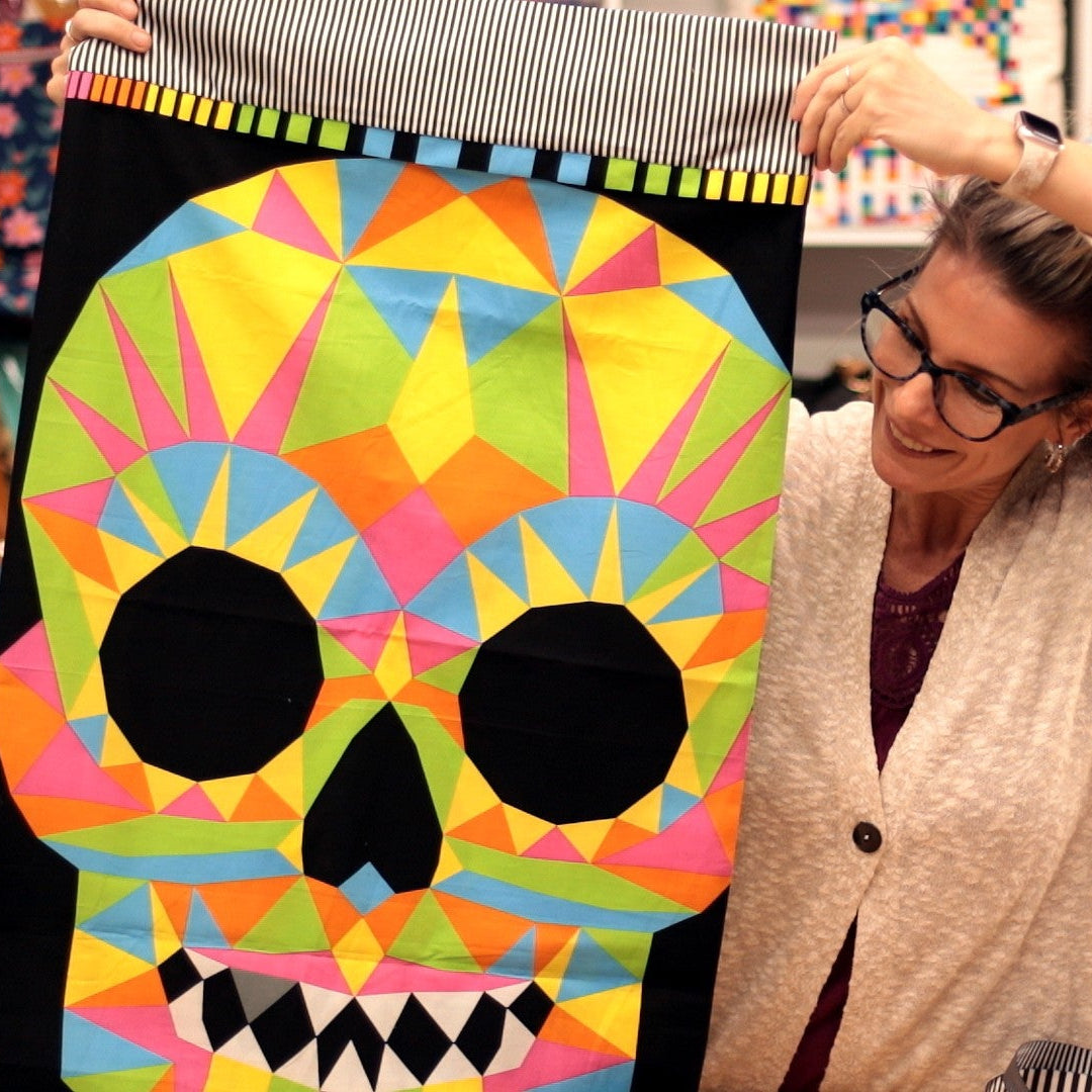 A woman in a cream cardigan and glasses holds up a large, colorful geometric sugar skull tapestry with a black background. The shop background features shelves filled with various patterned handbags.
