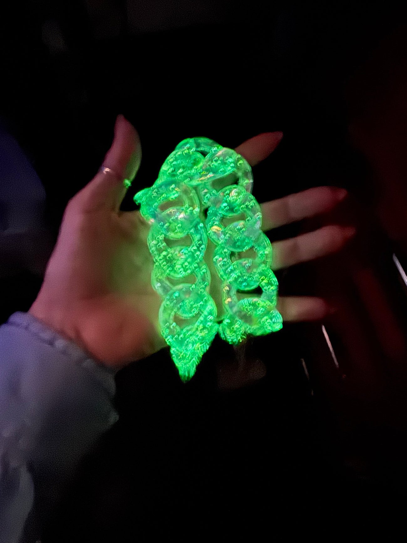 Hand holding a glowing green, UV-reactive clear acrylic chunky chain link accessory under a blacklight.