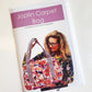 Front cover of a printed sewing pattern booklet titled "Joplin Carpet Bag" by Natalie Santini (@sewhungryhippie). The cover has a purple header band and shows a smiling woman with glasses and long blonde hair holding a large, colorful carpet-style bag. The bag features a vibrant, multi-colored floral pattern in pink, orange, and blue with thick, black-and-white striped Jacquard woven handles.