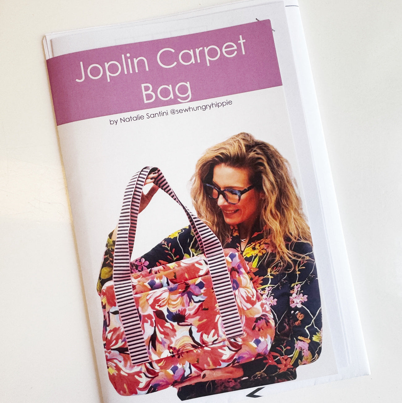 Front cover of a printed sewing pattern booklet titled "Joplin Carpet Bag" by Natalie Santini (@sewhungryhippie). The cover has a purple header band and shows a smiling woman with glasses and long blonde hair holding a large, colorful carpet-style bag. The bag features a vibrant, multi-colored floral pattern in pink, orange, and blue with thick, black-and-white striped Jacquard woven handles.