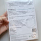 A hand holds a packaged sewing pattern for a 'Loopy Lou Bag' by Mrs H, showing the back of the clear plastic packaging with printed instructions, materials list, and a line drawing of the bag.