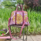 A larger magenta handbag with decorative floral pattern panels, resting on a stand outdoors with greenery in the background.