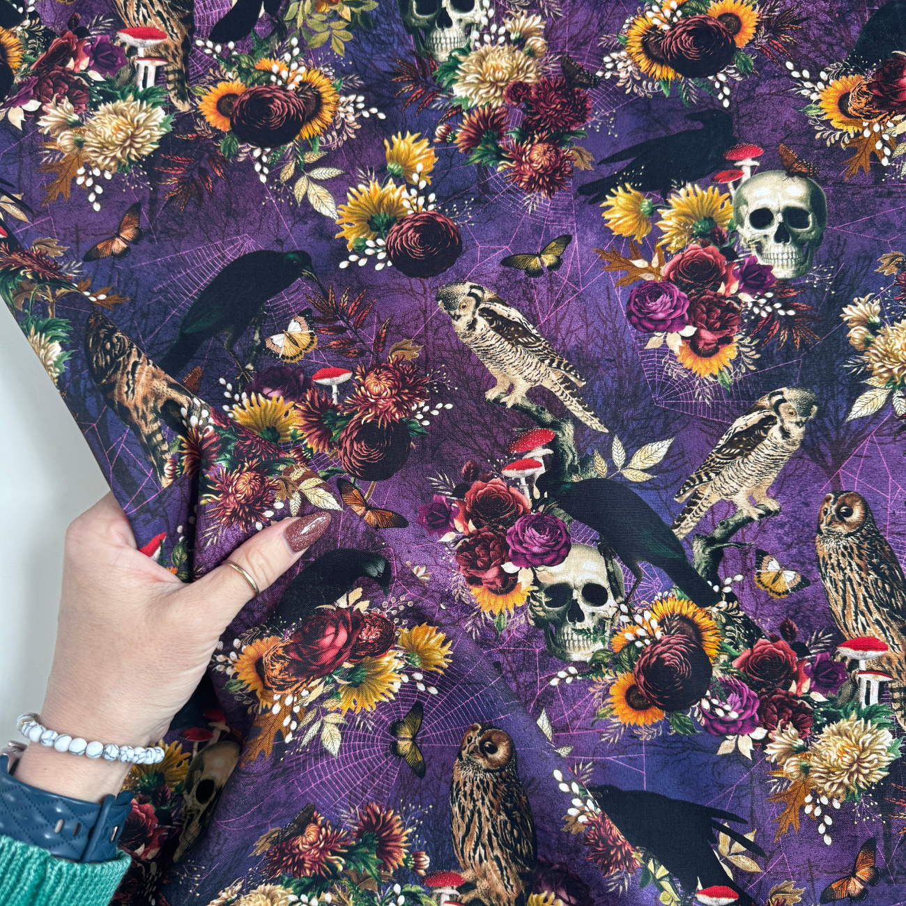 Hand holding a piece of Robert Kaufman Midnight Purple Dreadful Delights fabric to show the scale of the gothic skull, owl, and floral pattern.