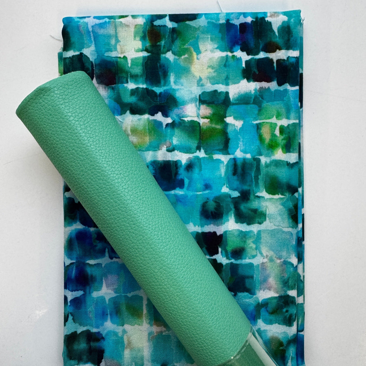 A green textured rolled vinyl placed diagonally over a folded piece of fabric with a blue, green, and teal watercolor pattern on a white background.