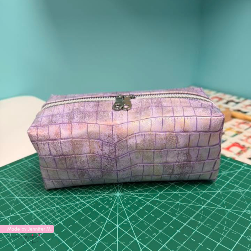 A rectangular zippered pouch with a purple and lavender textured grid pattern, resting on a green cutting mat with a teal background.