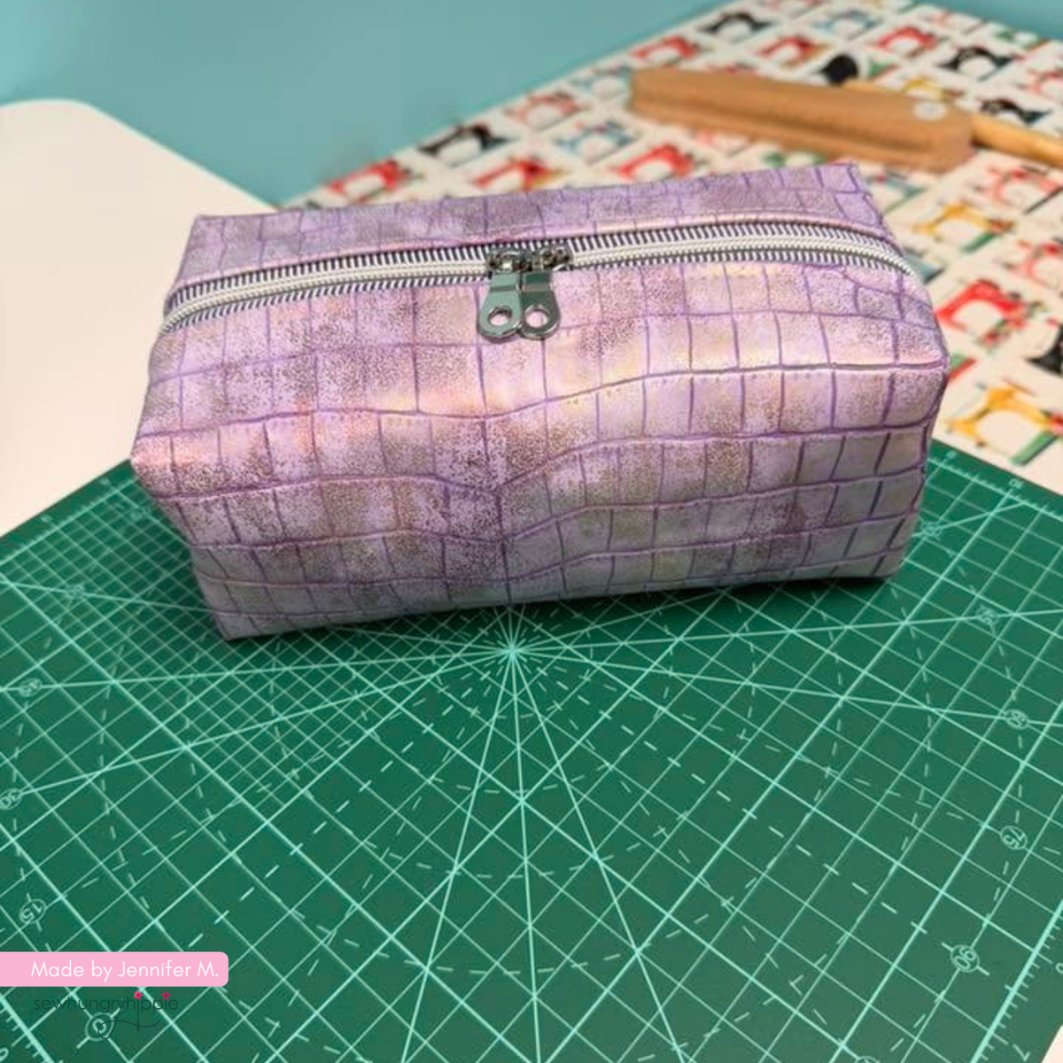 A rectangular zippered pouch with a purple and lavender crocodile-texture pattern, resting on a green cutting mat.