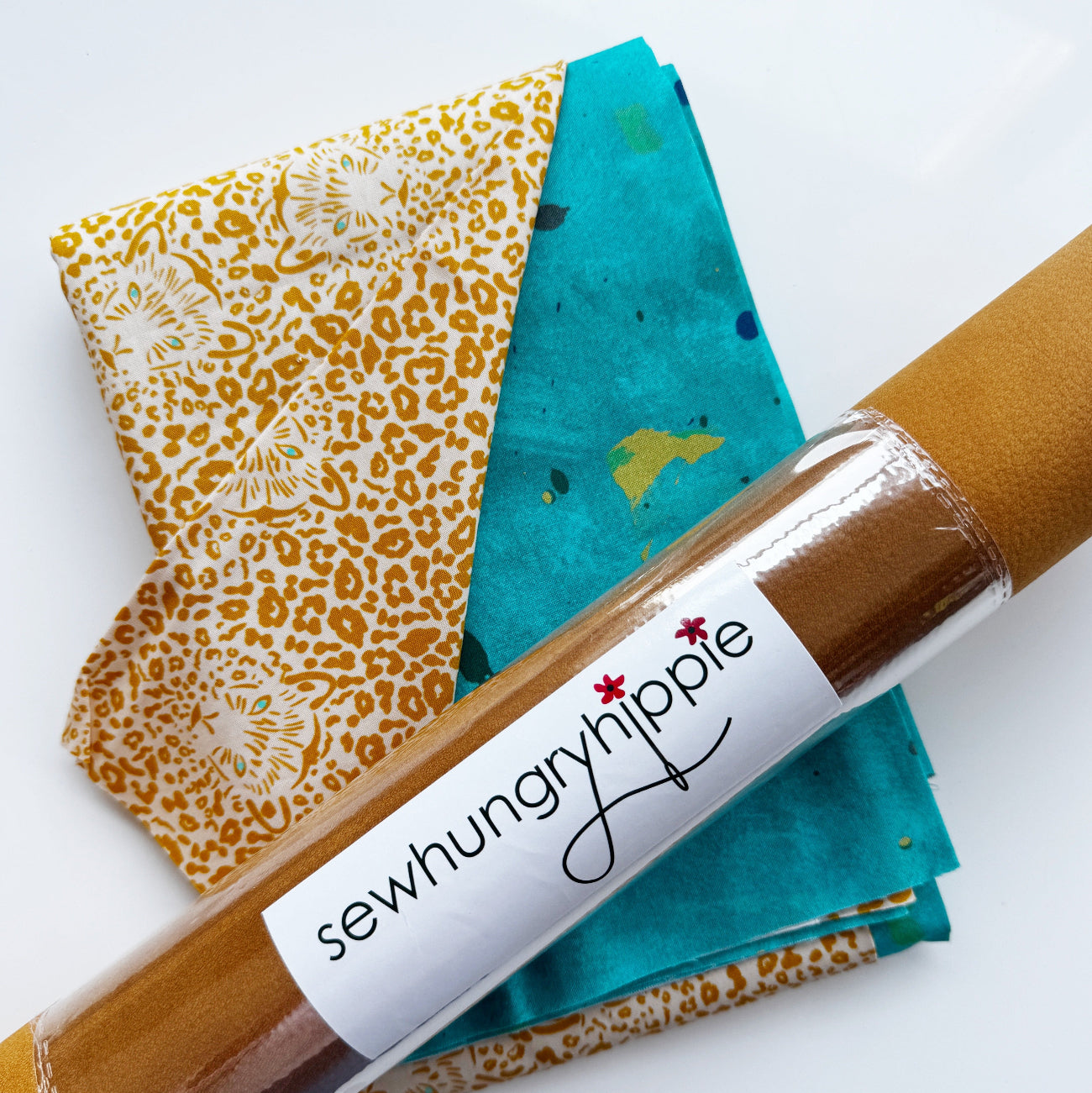 A flat-lay view of the craft materials for the Match Up #4 package, showing a roll of mustard-yellow faux suede with a "sewhungryhippie" label, alongside folded pieces of teal fabric and animal-print lining fabric.