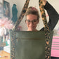 Natalie Santini holds up a large olive green quilted tote bag with a chevron pattern. She is holding the bag by its long leopard-print shoulder straps directly in front of her. The background shows a creative sewing studio with a sewing machine, a green dress form, and a pink neon "Peace Love Sewing" sign.