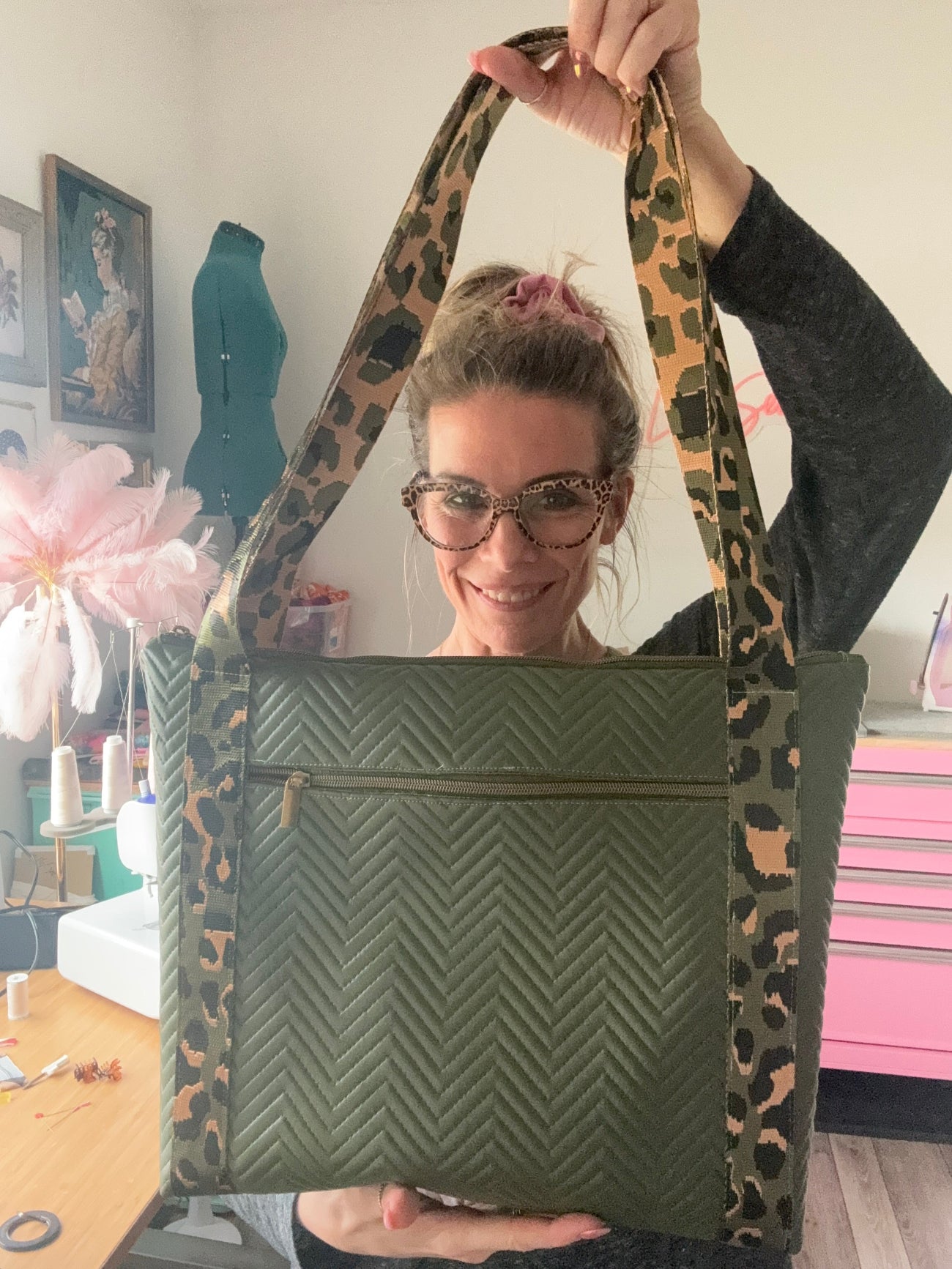Natalie Santini holds up a large olive green quilted tote bag with a chevron pattern. She is holding the bag by its long leopard-print shoulder straps directly in front of her. The background shows a creative sewing studio with a sewing machine, a green dress form, and a pink neon "Peace Love Sewing" sign.
