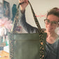 A woman with glasses smiles while holding up a green quilted chevron-pattern tote bag. She is positioned slightly to the right, lifting the leopard-print straps high with her right hand. The shot shows her sewing workspace, featuring a wooden table with tools, pink feathers, and a pink tool chest in the background.