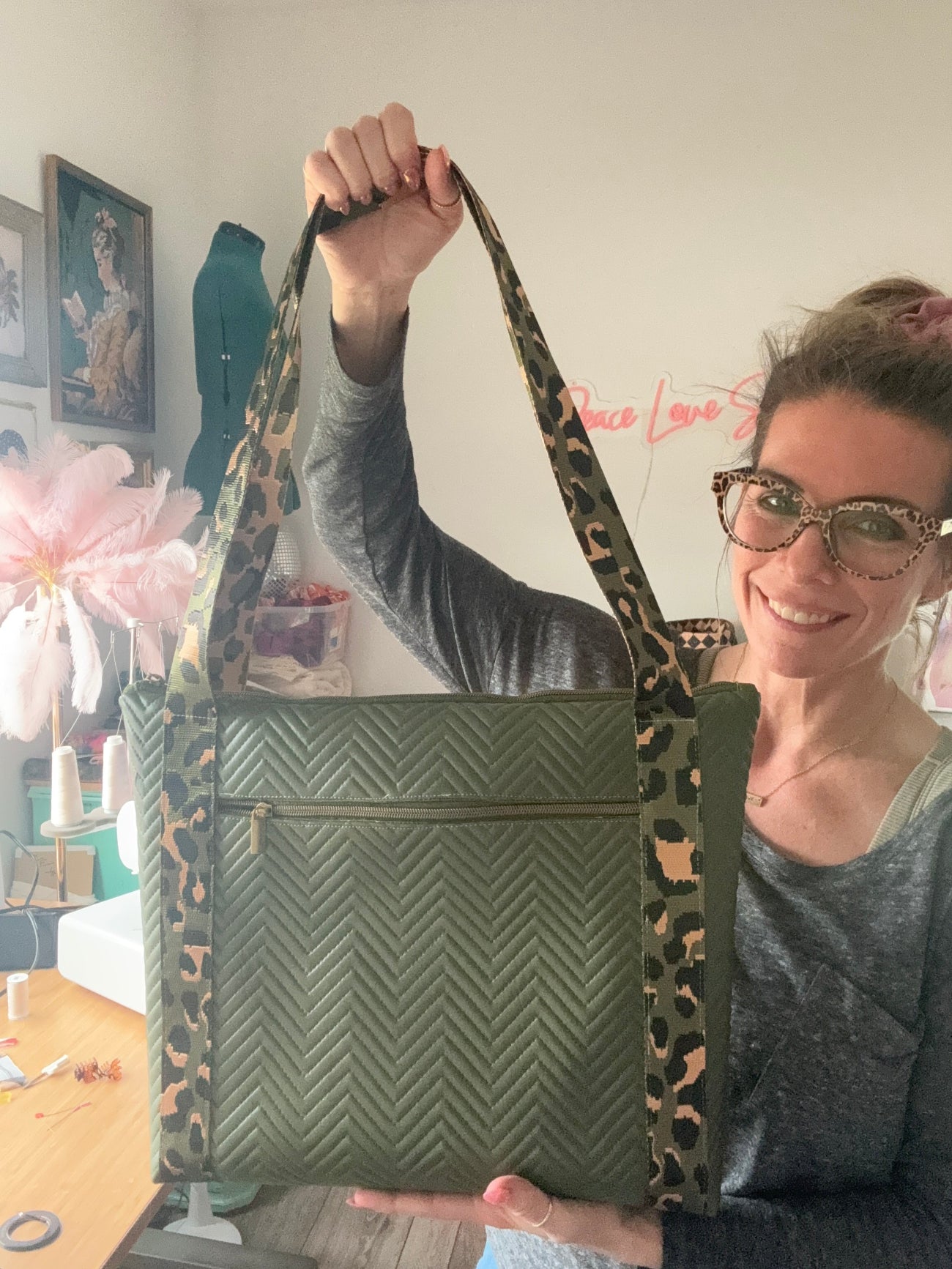 A woman with glasses smiles while holding up a green quilted chevron-pattern tote bag. She is positioned slightly to the right, lifting the leopard-print straps high with her right hand. The shot shows her sewing workspace, featuring a wooden table with tools, pink feathers, and a pink tool chest in the background.