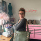 Natalie Santini smiling over her shoulder while wearing a large, olive green chevron-quilted tote bag on her shoulder. The bag features thick leopard-print straps. She is standing in a craft room with a Juki sewing machine on the left and a pink tool chest underneath a neon "Peace Love Sewing" sign on the wall.
