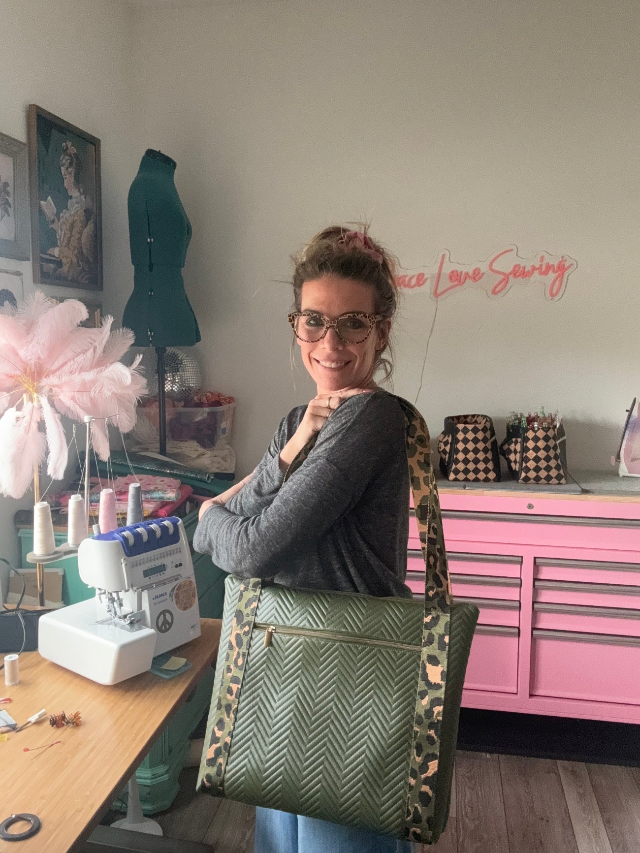Natalie Santini smiling over her shoulder while wearing a large, olive green chevron-quilted tote bag on her shoulder. The bag features thick leopard-print straps. She is standing in a craft room with a Juki sewing machine on the left and a pink tool chest underneath a neon "Peace Love Sewing" sign on the wall.