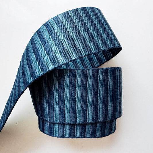 A close-up shot of a 2-inch wide, heavy-duty woven fabric webbing strap on a white background. The strap features horizontal stripes alternating between dark navy blue and a lighter slate blue. The webbing is folded into a loose, architectural loop, highlighting its thick, ribbed tactical texture ideal for durable bag straps, tote handles, and backpacks.