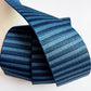 An overhead product view of a 2-inch wide striped woven webbing strap against a solid white surface. The strap features a repeating vertical pinstripe pattern in alternating shades of deep navy blue and steel blue. The material is neatly wrapped into a cylindrical roll with the top end arching up and over, displaying the smooth, sturdy weave on both sides of the strap.