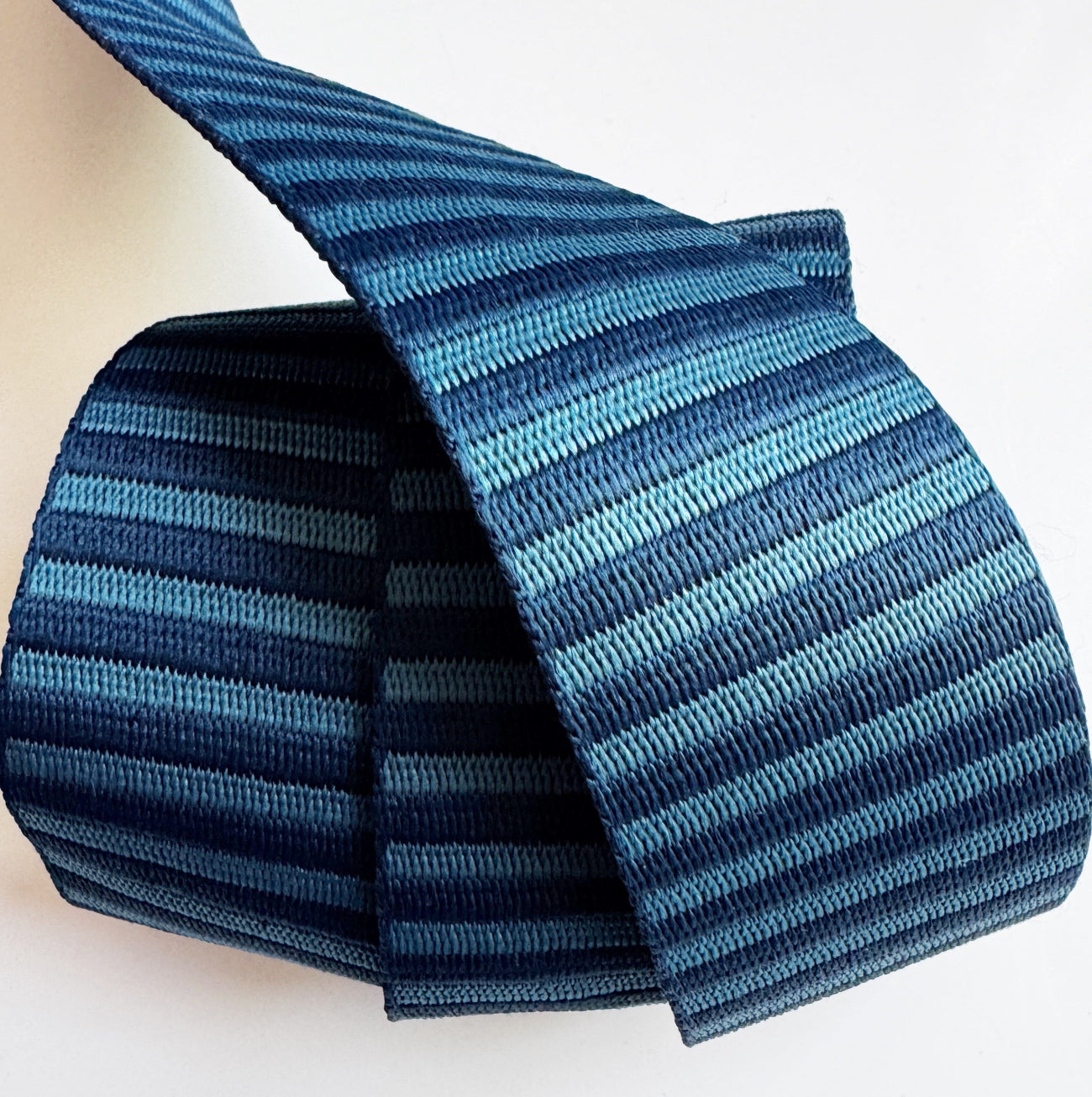An overhead product view of a 2-inch wide striped woven webbing strap against a solid white surface. The strap features a repeating vertical pinstripe pattern in alternating shades of deep navy blue and steel blue. The material is neatly wrapped into a cylindrical roll with the top end arching up and over, displaying the smooth, sturdy weave on both sides of the strap.
