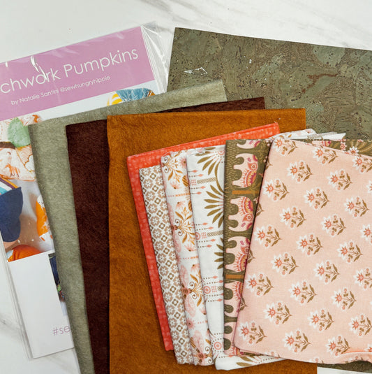 A stack of fabric swatches in warm autumn and pumpkin-themed patterns, including solid rust, mustard, olive green, cream with floral motifs, and light pink with small floral prints. A package labeled 'Swatch Pumpkins' by Natalie Sandoval is partially visible underneath.