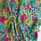 A piece of premium 100% cotton sewing fabric by the yard featuring a vibrant, maximalist botanical print designed by Sally Kelly for Windham Fabrics. The intricately detailed, painterly pattern showcases large stylized flowers and lush garden foliage in bright shades of magenta pink, sunny yellow, teal blue, lime green, and coral over a clean white background. The fabric is casually gathered and draped to highlight its soft texture and fluid drape.