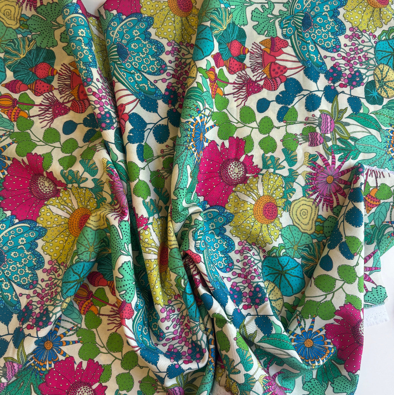 A piece of premium 100% cotton sewing fabric by the yard featuring a vibrant, maximalist botanical print designed by Sally Kelly for Windham Fabrics. The intricately detailed, painterly pattern showcases large stylized flowers and lush garden foliage in bright shades of magenta pink, sunny yellow, teal blue, lime green, and coral over a clean white background. The fabric is casually gathered and draped to highlight its soft texture and fluid drape.