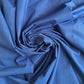 A close-up, top-down view of the royal blue nylon bag lining fabric, artistically arranged in a tight swirl or rosette shape. The lighting highlights the fine, woven, water-resistant texture of the material along the edges of its deep, dramatic folds.