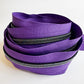 A loose, nested pile of continuous zipper tape on a plain white background. The fabric tape is a vibrant grape purple, and the heavy-duty nylon coil teeth feature a dark gunmetal finish with a subtle rainbow iridescent shimmer that catches the light.