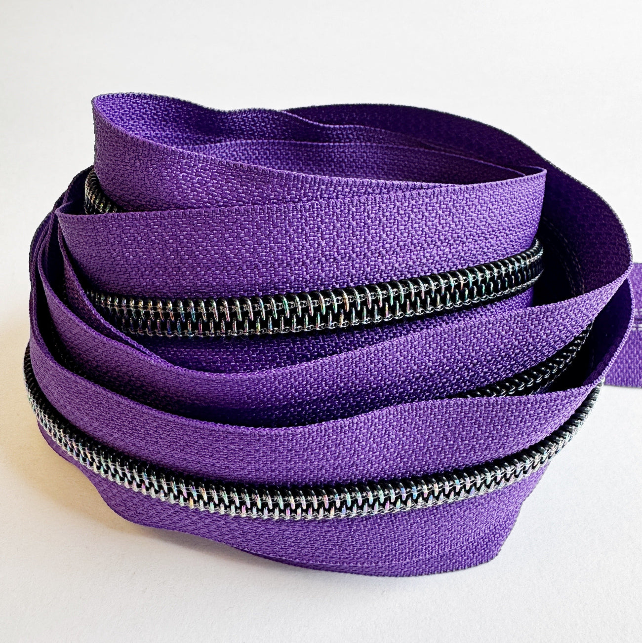 A loose, nested pile of continuous zipper tape on a plain white background. The fabric tape is a vibrant grape purple, and the heavy-duty nylon coil teeth feature a dark gunmetal finish with a subtle rainbow iridescent shimmer that catches the light.