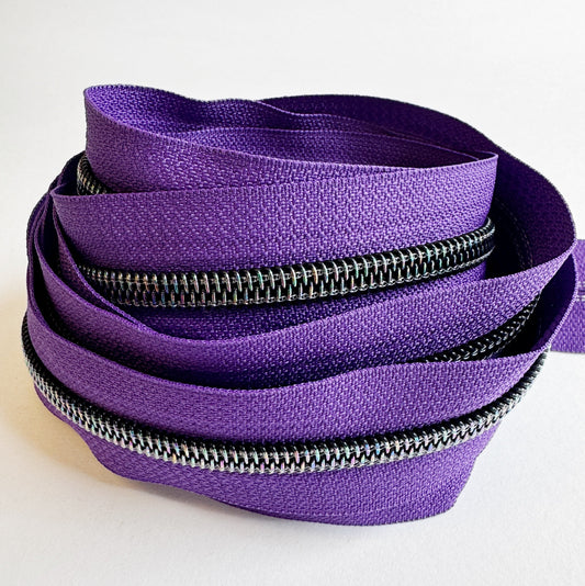 A loose, nested pile of continuous zipper tape on a plain white background. The fabric tape is a vibrant grape purple, and the heavy-duty nylon coil teeth feature a dark gunmetal finish with a subtle rainbow iridescent shimmer that catches the light.