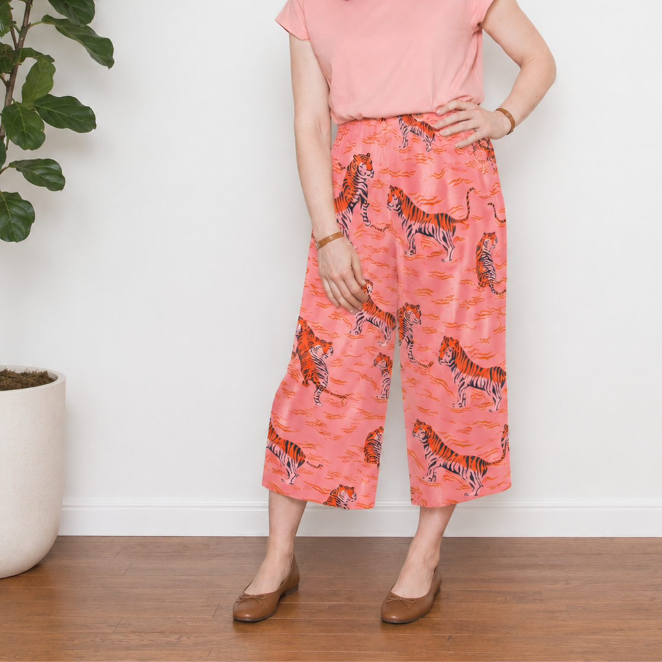 A person standing from the neck down modeling wide-leg cropped pants made from Ruby Star Society Poolside Too Rayon Fabric in Sorbet (Pink). The pants feature a repeating orange and white swimming tiger pattern on a pink background, paired with a solid pink t-shirt and brown ballet flats.