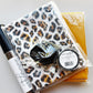 A sewing kit bundle containing a leopard print fabric package, a black sewing machine needle, a black elastic loop, and a yellow mesh bag, all in clear plastic wrapping with a SewhungryNipple brand label visible.